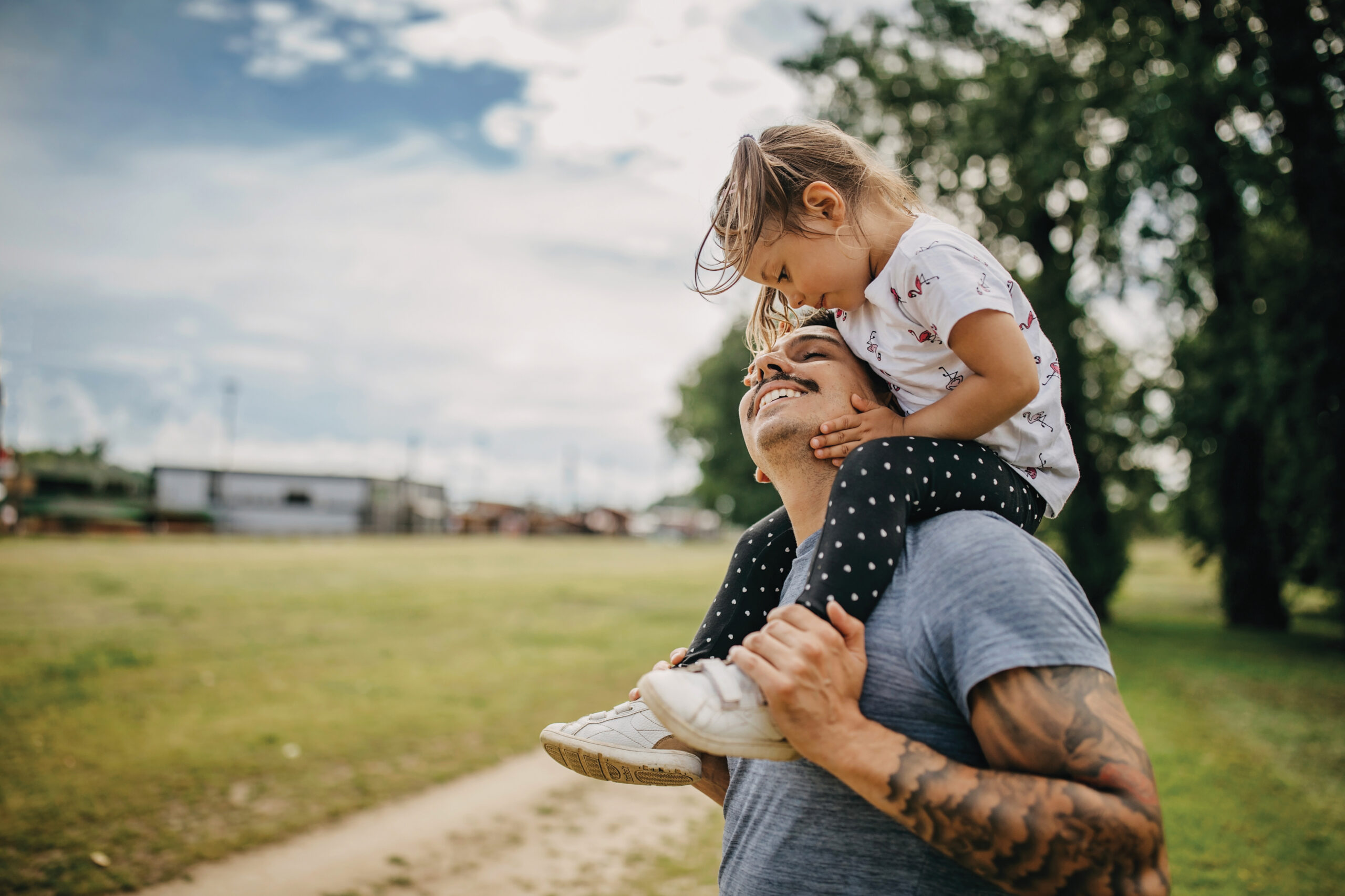 A smiling man with a tattooed arm carries a young girl on his shoulders in a grassy outdoor park. The girl leans down, touching his face affectionately as they both smile. Trees and a blue sky with clouds are visible in the background.