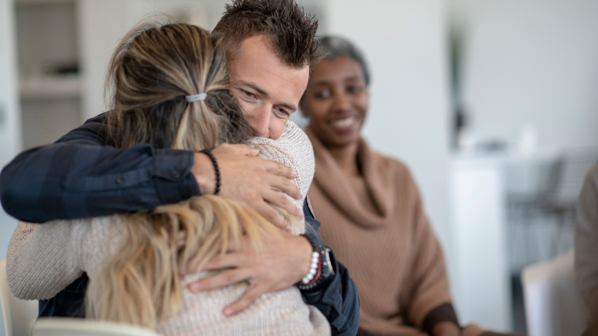 A heartfelt embrace between two individuals in a supportive environment, conveying hope and the compassionate care provided by addiction recovery services, with a professional looking on with an encouraging smile.