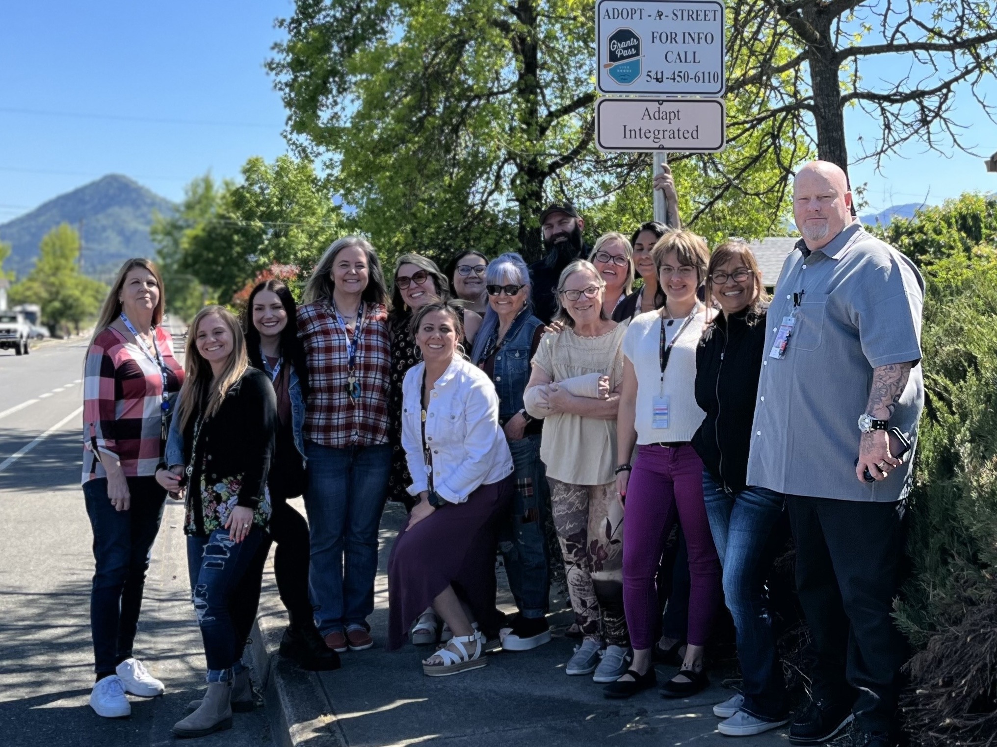 Group Photo at Adopt-a-Street Sign: A group of Adapt Integrated Health Care employees stands smiling under an Adopt-a-Street sign on a sunny day, with a scenic mountain in the background.