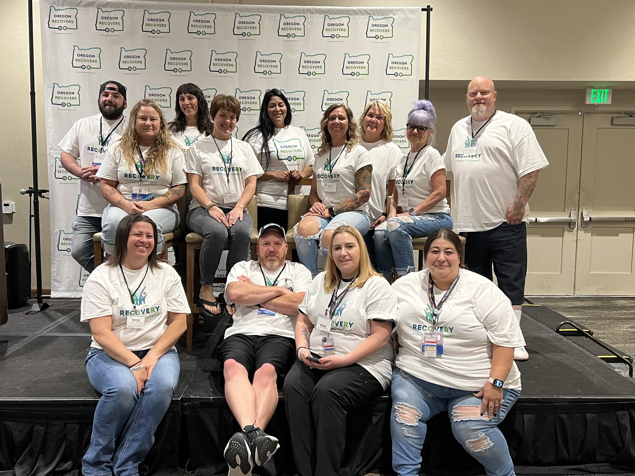 Outreach Peers: A group of Adapt Integrated Health Care outreach peers, wearing matching T-shirts, posing together on a stage with a banner in the background.