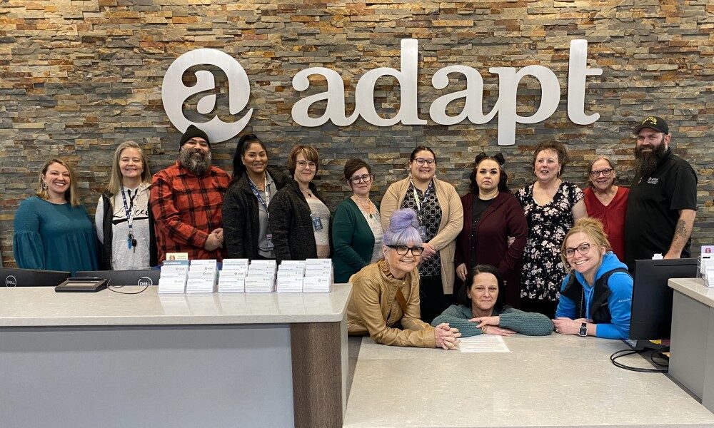 A diverse group of Adapt Integrated Health Care staff smiling at the reception desk, with the Adapt logo on a stone wall background, symbolizing welcoming and professional service in Southwest Oregon.