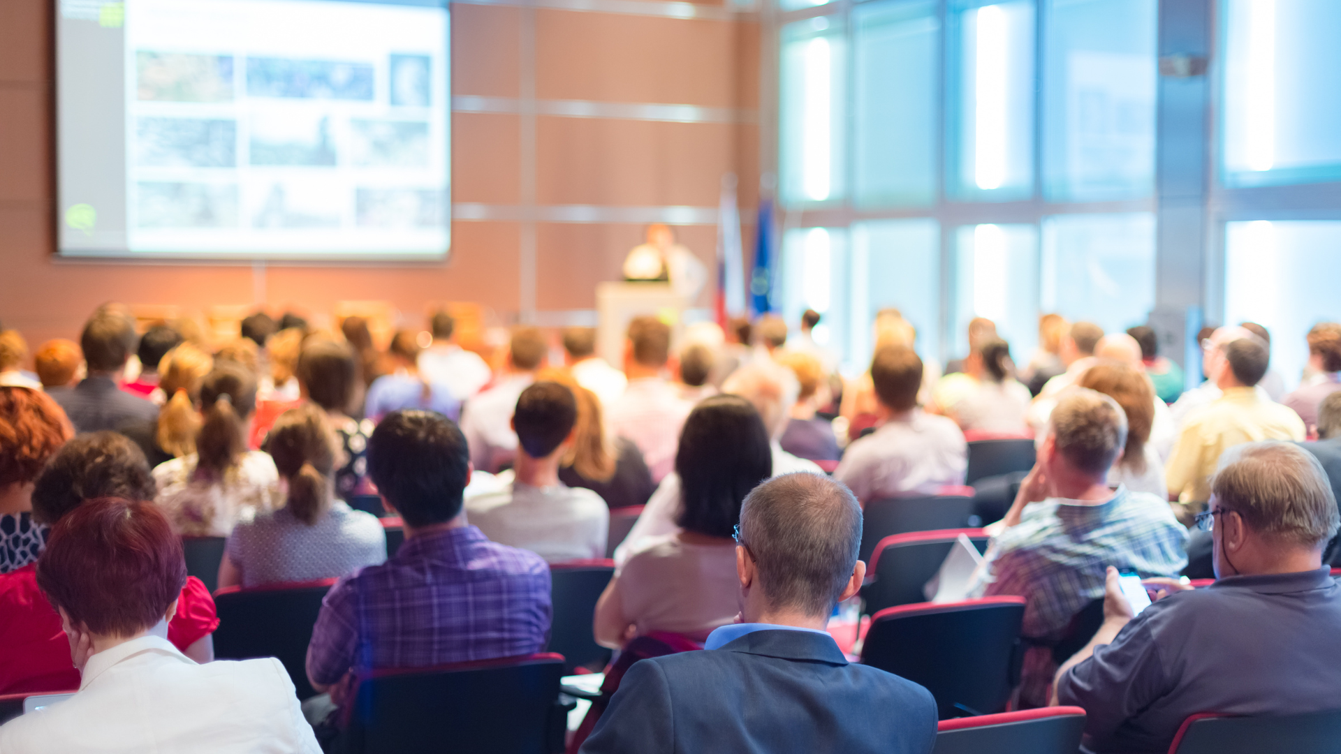 Audience attending a presentation in a large conference hall, viewed from the back, focusing on the crowd and a presenter in the distance