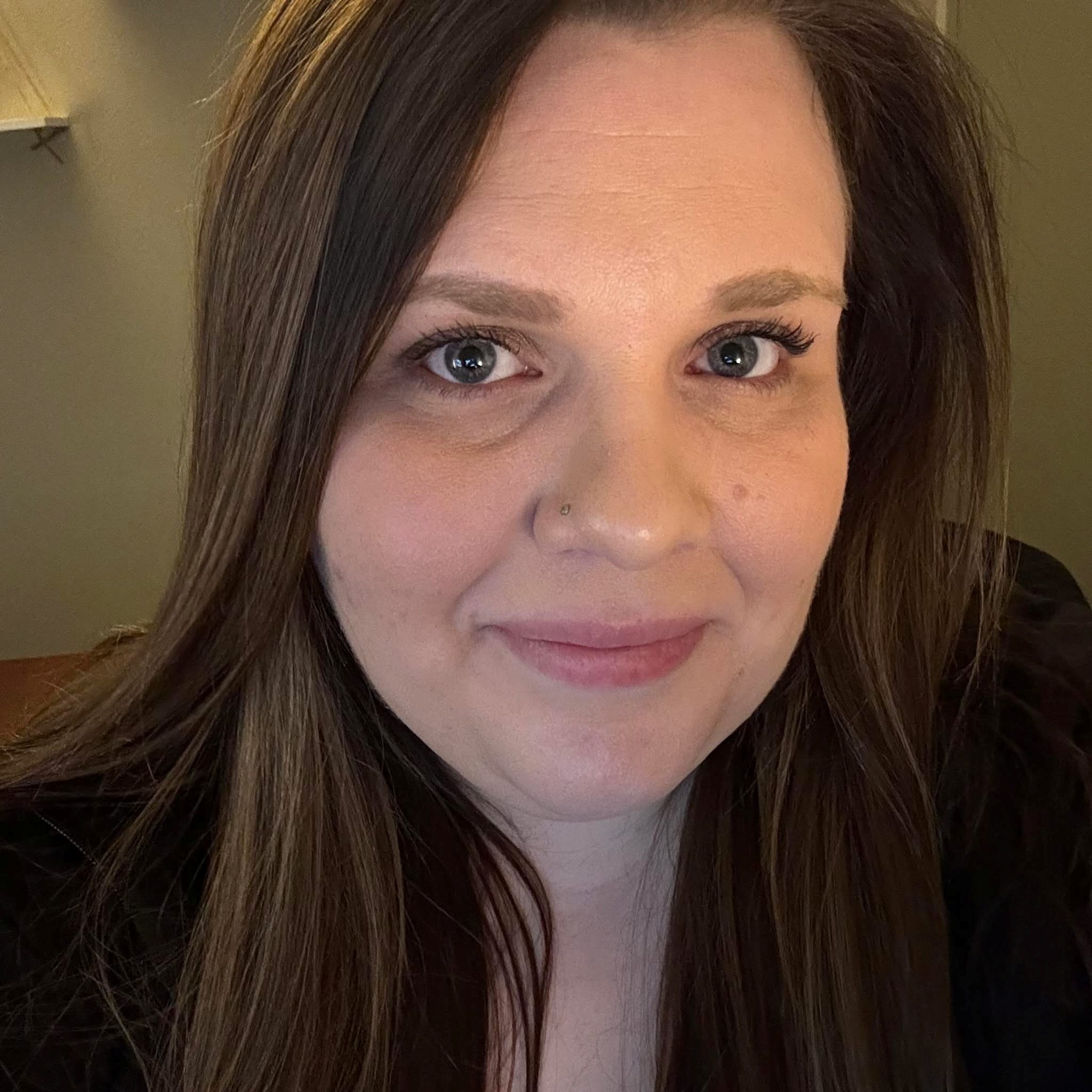 A close-up headshot of a woman with long, straight brown hair smiling softly at the camera. She has a small nose piercing and is wearing a black top. The background is softly lit and neutral.